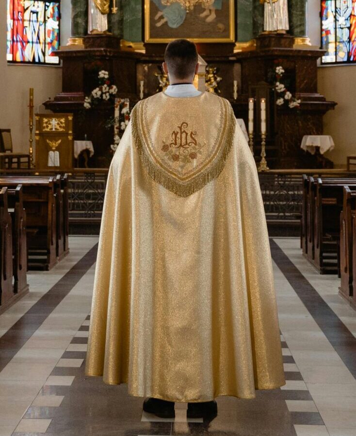 A priest in a golden robe walks down the aisle of a grand cathedral with stained glass windows.