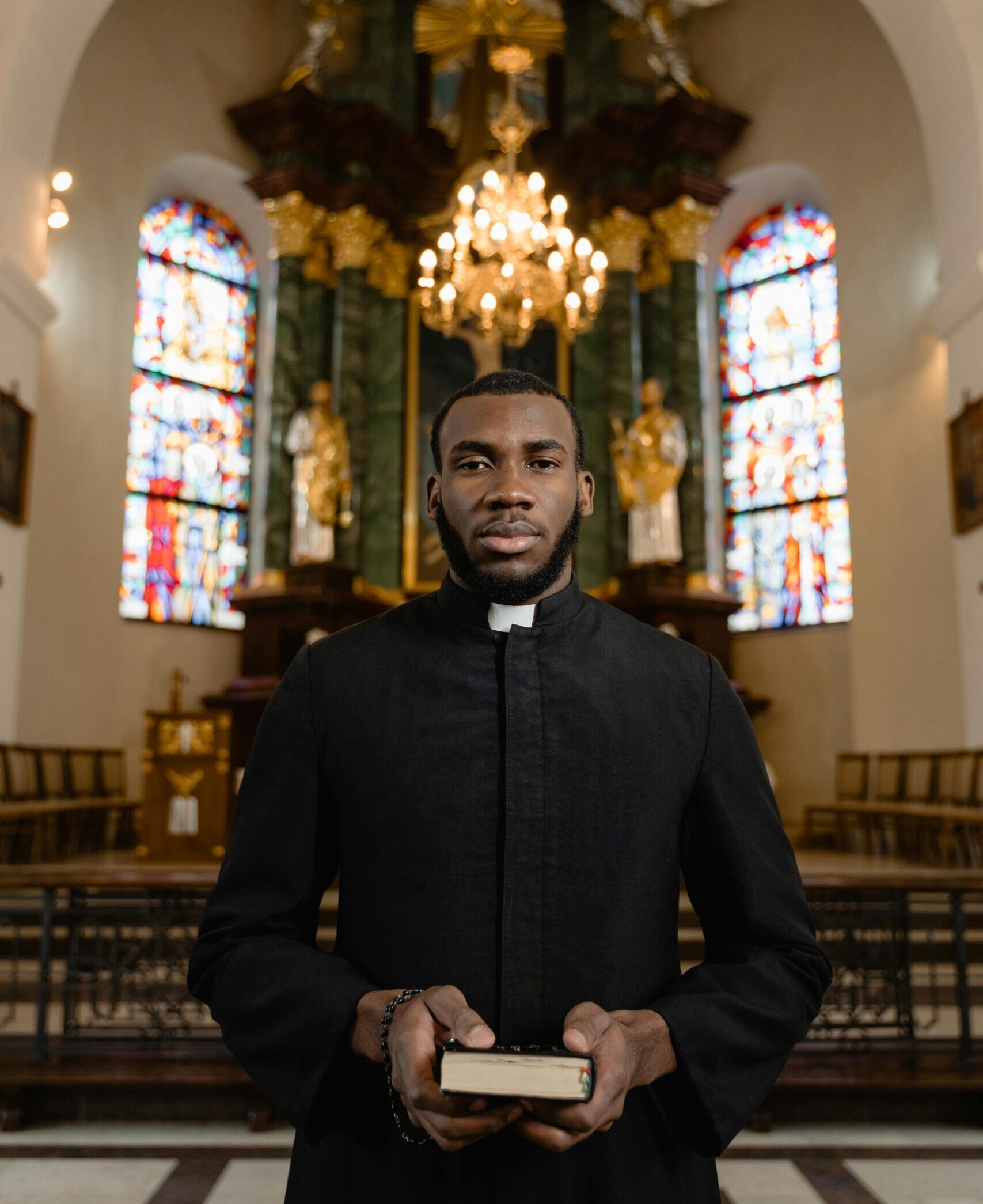 A priest holds a Bible inside a beautifully decorated cathedral with stained glass windows.